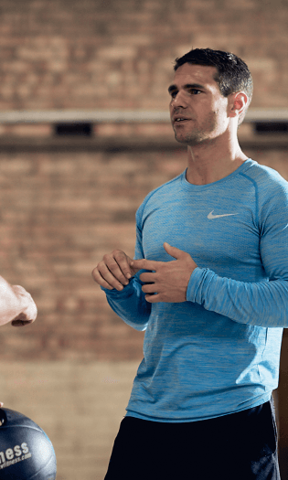A man in a blue Nike long-sleeve shirt gesturing while speaking, standing in front of a brick wall.