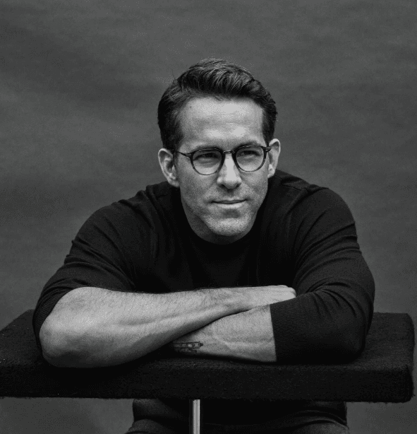 A black and white portrait of a man with short dark hair wearing glasses and a black sweater, resting his arms on a surface.